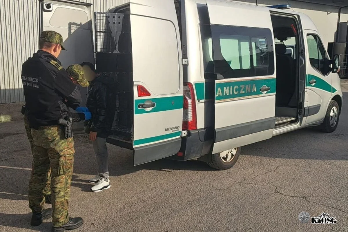 Funkcjonariusze Straży Granicznej przy białym furgonie z napisem „GRANICZNA” zatrzymują młodego mężczyznę na parkingu przed budynkiem.