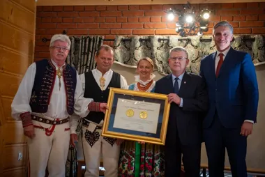 Grupa osób, w tym trzy w strojach ludowych i dwie w garniturach, wręcza lub odbiera uroczystą dyplomową ramę w pomieszczeniu z ceglanym kominkiem i drewnianymi