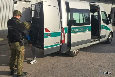 Funkcjonariusze Straży Granicznej przy białym furgonie z napisem „GRANICZNA” zatrzymują młodego mężczyznę na parkingu przed budynkiem.