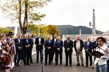 Grupa mężczyzn w garniturach i osoby w strojach ludowych stoją na moście podczas uroczystości, podczas gdy kobieta w białej kamizelce przemawia do mikrofonu.