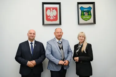 Trójka osób w eleganckich strojach stoi przed białą ścianą z dwoma herbami w ramkach: polskim orłem i herbem Orawy.