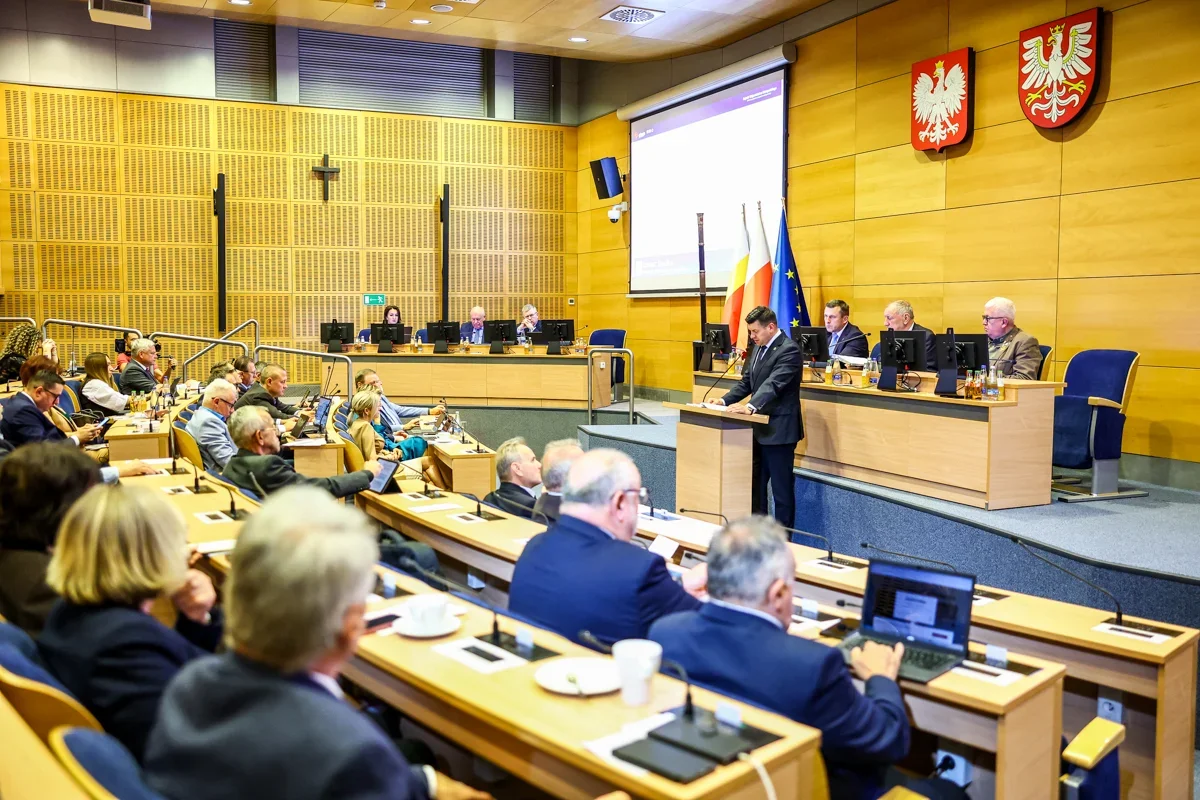 Mężczyzna w garniturze przemawia z mównicy w sali posiedzeń z drewnianymi panelami, przed siedzącymi uczestnikami i osobami przy stole prezydialnym, z polskimi